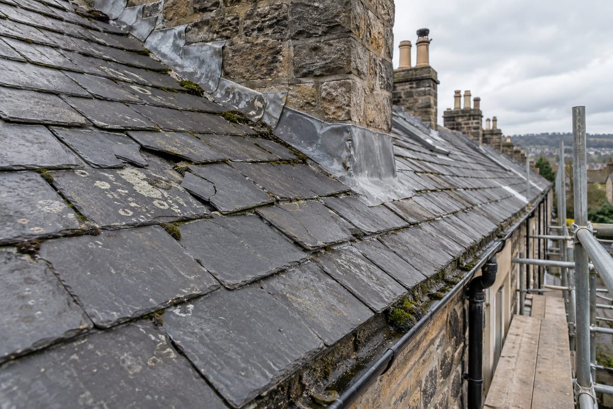 Older terrace slate roof with cracked and slipped slates, moss in the joints, lifted lead soakers at the stone chimney and a blocked gutter